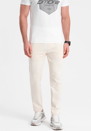 Beige bavlněné kalhoty s rovný střih, s bočními kapsami, kombinované s bílým grafickým tričkem a šedými teniskami. Jednoduchý design.