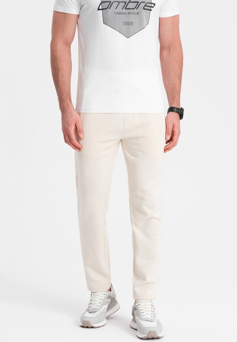 Beige Baumwollhose mit geradem Schnitt und seitlichen Taschen, kombiniert mit einem weißen Grafik-T-Shirt und grauen Sneakern. Schlichtes Design.