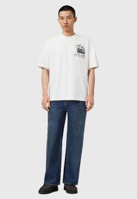 Tricou alb din bumbac cu mâneci scurte, având o grafică imprimată pe piept. Asociat cu blugi largi de culoare albastră închis.