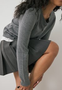 Donna con capelli ricci sistemando una scarpa nera, indossando un cardigan grigio, una gonna plissettata e una camicia bianca su uno sfondo semplice.