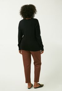 Sort striktrøje med en afslappet pasform, parret med brune slim-fit bukser. Sorte flade sko med guldnitter på tåen. Neutral baggrund.