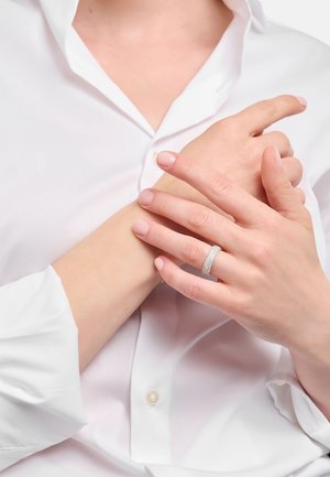 Anello d'argento con più file di piccole gemme bianche, indossato su una mano con unghie naturali, su uno sfondo di camicia bianca.