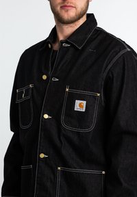 Chaqueta de trabajo de denim negro con herrajes dorados, dos bolsillos en el pecho y costuras en blanco en contraste. Presenta un parche cuadrado del logo en el bolsillo izquierdo.