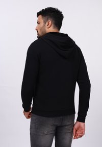 Schwarzer Kapuzenpullover mit langen Ärmeln, gerippten Bündchen und Saum sowie weicher Textur. Getragen mit grauen Jeans, von hinten betrachtet.