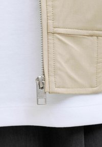 Detail béžovej látky so strieborným zipsom a našitým vreckom vedľa bieleho a čierneho vrstveného oblečenia.