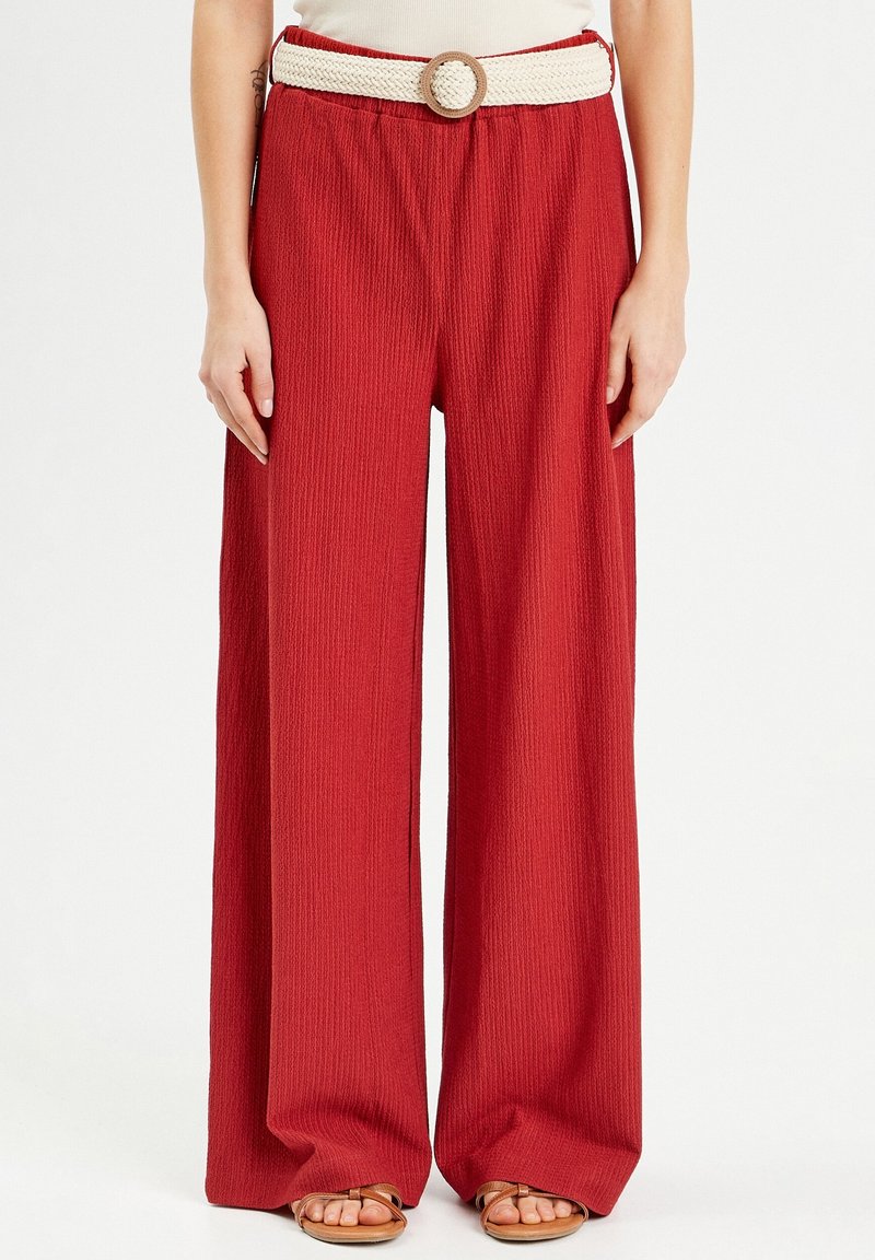 Pantalon large rouge texturé avec une ceinture tissée blanche et une boucle circulaire, porté avec des sandales ouvertes beige sur un fond clair.