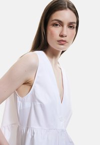 Vestido blanco sin mangas con un escote en V profundo, que presenta un detalle de botones y un dobladillo acampanado, fabricado con una tela ligera de textura suave.