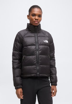 Frau trägt eine schwarze Steppjacke mit Frontreißverschluss und Logo auf der Brust, steht vor einem einfarbigen hellen Hintergrund.