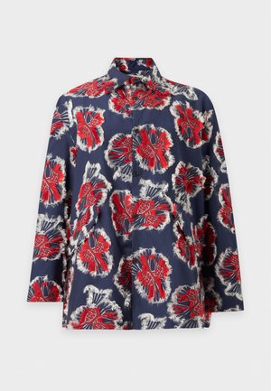 Une femme porte un chemisier bleu marine à motif floral rouge, un pantalon ample marron, des chaussures noires et un sac à main marron devant un fond uni.
