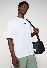 T-shirt blanche à manches courtes avec logo Adidas noir, associée à un short vert clair. Sac de sport noir avec détails en maille tenu par le modèle.