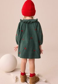 Groene corduroy jurk met rode kersenborduring, gerimpelde kraag en lange mouwen. Gecombineerd met rode sokken en lichtbruine laarzen. Rode gebreide muts op het hoofd.