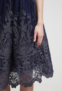 Laivastonsininen mekko, jossa on monimutkaisia hopeakirjailtuja kukkakuoseja, läpikuultava kerroksellinen rakenne ja aaltoileva helma. Kädentiedot näkyvät reunassa.