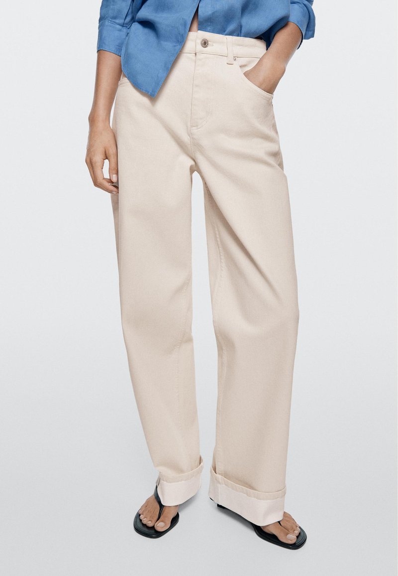 Donna che indossa pantaloni beige a gamba larga con risvolti, una camicia blu con maniche risvoltate e sandali infradito neri, in piedi contro uno sfondo semplice.