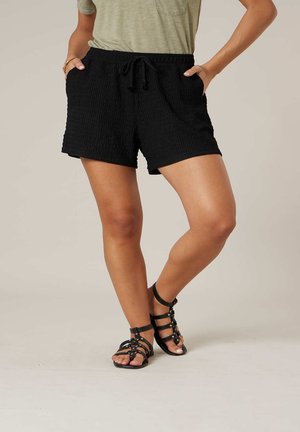 Femme portant un short noir texturé à cordon de serrage et des sandales noires à lanières, mains dans les poches, debout avec une jambe croisée sur l'autre.