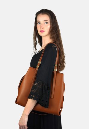 Brown leather tote bag with structured shape, gold hardware accents, and side cutouts. The bag features two long shoulder straps.