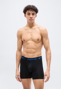 Νέος άνδρας με σγουρά μαλλιά που φοράει μαύρα εσώρου χα μπόξερ Nike με μπλε λογότυπο, στεκόμενος μπροστά σε ένα απλό λευκό φόντο.