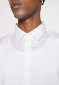 Biała koszula zapinana na guziki z teksturowanej tkaniny, z ostrym kołnierzykiem i przezroczystymi guzikami, ukazująca część szyi i ramienia.