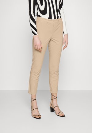 Personne portant un pantalon beige slim, des sandales à talons noires à brides et un haut à manches longues noir et blanc à motifs, debout devant un fond uni.