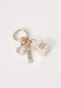Arany fém kulcstartó, amelyen lakat, "GUESS" feliratú címke és "G" betű található. Simább textúra, fényes felülettel és gravírozott részletekkel.