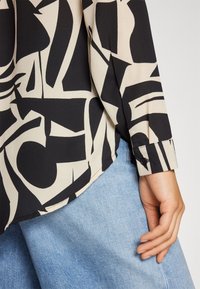 Blusa a maniche lunghe nera e crema con motivo, caratterizzata da polsini con bottone e orlo curvato, abbinata a jeans in denim chiaro.