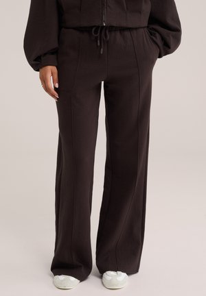 Personne portant un pantalon noir ample avec des coutures à l'avant et des chaussures blanches décontractées, debout avec une main dans la poche sur fond uni.