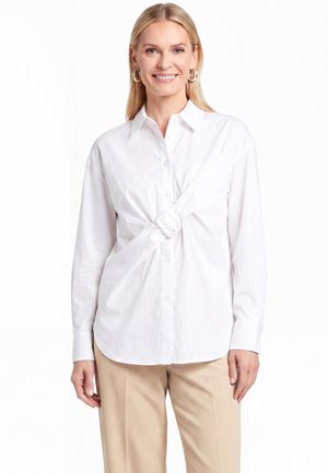 Lächelnde Frau mit langen blonden Haaren, die ein weißes Hemd mit Knöpfen und vorderer Schnalledetail sowie beige Hosen vor weißem Hintergrund trägt.