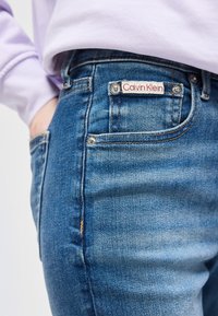 Κοντινή λήψη τσέπης από μπλε τζιν με ετικέτα Calvin Klein και ένα χέρι που μπαίνει μέσα στην πίσω τσέπη, φορώντας ανοιχτό μωβ μπλουζάκι.