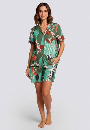 Woman standing barefoot wearing green tropical print short-sleeve button-up shirt and matching shorts with hand in pocket.