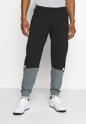 Man draagt zwart-grijze joggerbroek met blokkleuren en witte Nike-sneakers, staand tegen een witte achtergrond.