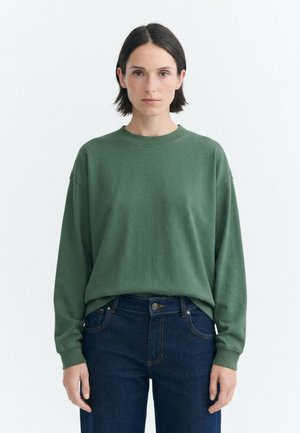 Donna con capelli corti e scuri, che indossa una camicia verde a maniche lunghe e jeans blu scuro, in piedi davanti a uno sfondo bianco semplice.