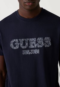 Marineblauwe katoenen T-shirt met witte decoratieve "GUESS" tekst in een patroonlettertype, korte mouwen en een ronde hals.