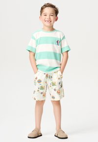 Gestreiftes T-Shirt in Minzgrün und Creme mit einem Kaktus-Patch, kombiniert mit cremefarbenen Shorts mit bunten Druckmotiven. Helle Slipper.