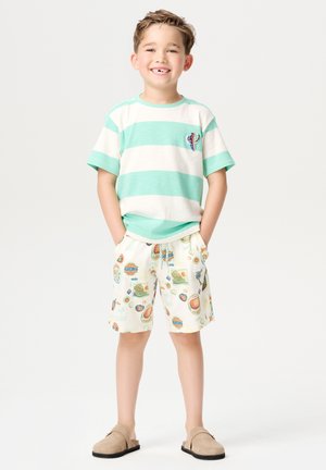 Gestreiftes T-Shirt in Minzgrün und Creme mit einem Kaktus-Patch, kombiniert mit cremefarbenen Shorts mit bunten Druckmotiven. Helle Slipper.