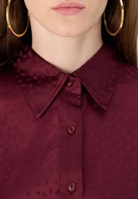 Camisa bordeaux de botão com colarinho pontudo, apresentando um padrão de folhas subtil, textura suave e botões coordenados. Brincos grandes em forma de argola visíveis.