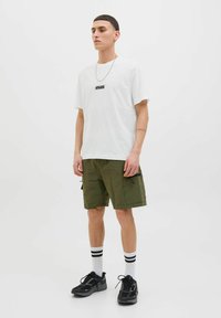 White cotton t-shirt with black logo, paired with olive green cargo shorts featuring side pockets, black accents, and black athletic shoes with white socks.