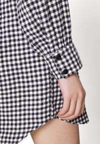 Camisa de cuadros en blanco y negro con una textura suave, que presenta un puño abotonado y un dobladillo ligeramente curvado. Mano visible sosteniendo la tela.