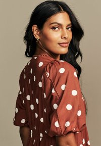 Ruskea pilkullinen mekko, jossa on puffihihat ja joustava vyötärö. Sisältää valkoisia pisteitä ja hienovaraisia ompeleita kaula-aukon ja hihansuiden reunoilla.