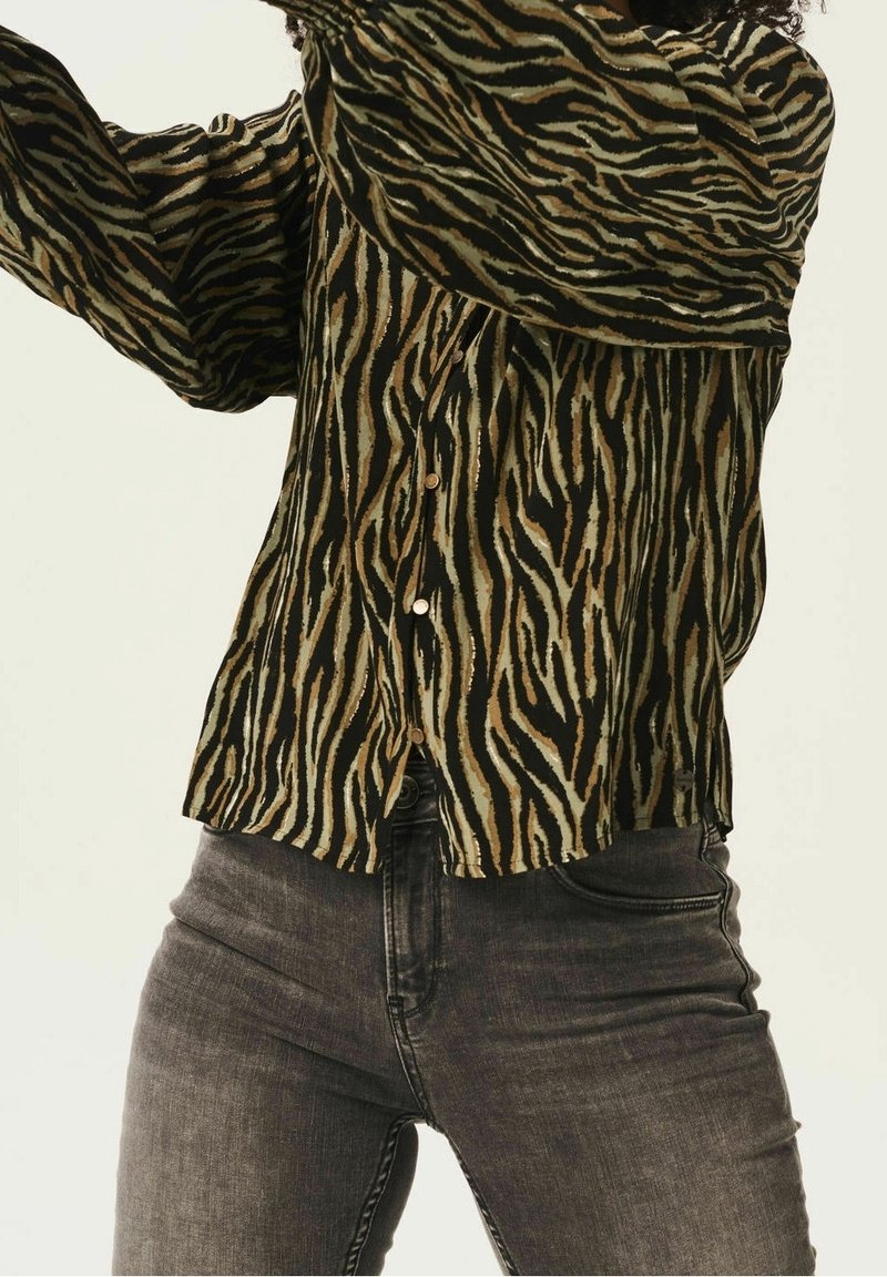 Personne portant un t-shirt à manches longues rayé tigre et un jean noir délavé, les bras levés et le torse visible, sur un fond uni.