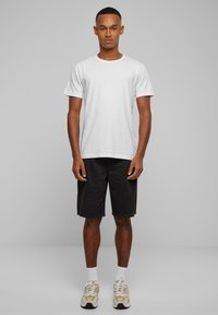 Tricou alb din bumbac, pantaloni scurți negri, șosete albe și adidași bej cu accente închise. Modelul stă pe un fundal gri simplu.