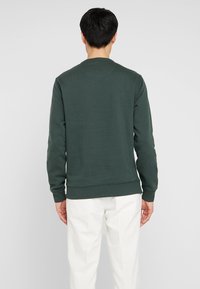 Ciemnozielony sweter z okrągłym dekoltem, prążkowanymi mankietami i dolnym wykończeniem. Gładka tekstura, prosty design i komfortowe dopasowanie do białych spodni.