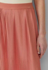 Plisségroen rok van koraalstof met een gladde textuur. De tailleband is elastisch en er is een klein labeldetail aan de bovenkant.