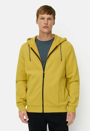 Jonge man die een gele hoodie met rits draagt over een blauw overhemd en zwarte broek, staand tegen een eenvoudige witte achtergrond.