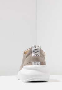 Szary zamszowy trampki z białą podeszwą, z wyraźną tylną pętlą z czarnym napisem. Gładka powierzchnia i zaokrąglony kształt.