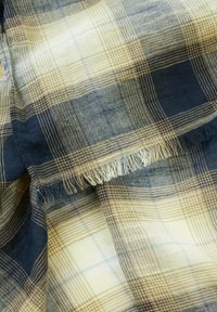 Vævet stof i blå, beige og creme farvet plaid-mønster med frynsede kanter; glat tekstur og varierede gitterlinjer gennem hele materialet.