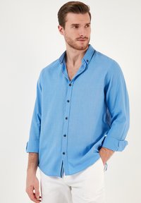 Camisa de botones azul claro hecha de una tela suave, con cuello y botones oscuros. Las mangas están ligeramente remangadas.