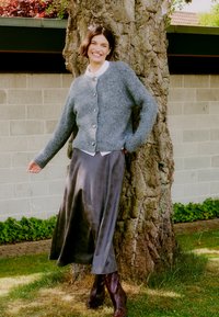 Gilet en tricot gris avec de grands boutons, associé à une jupe sombre en soie et des bottes marron montantes, posé contre un tronc d'arbre.