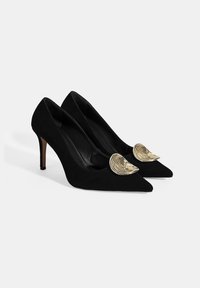 Escarpins noirs en daim avec des pointes et un accent en spirale doré à l'avant. Les talons sont fins et mesurent environ 7,5 cm.