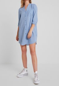 Abito a righe blu e bianche con bottoni, colletto e una tasca sul petto. Presenta maniche lunghe arrotolate e una vestibilità comoda. Indossato con sneakers bianche.