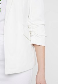 White blazer with a smooth texture, featuring a relaxed fit and gathered sleeve details; pair of side pockets on front.