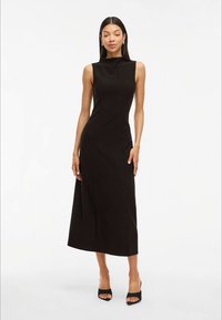 Vestido midi preto sem mangas com decote alto, corpete ajustado e saia reta. Tecido suave e costuras mínimas. Sapatos de salto aberto pretos.